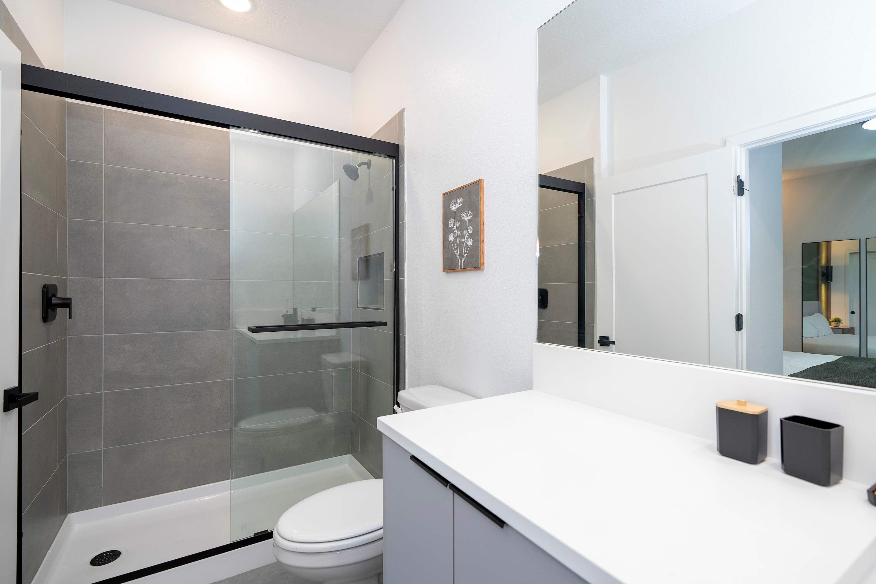 A modern bathroom with a glass shower enclosure, a white toilet, and a white vanity with two black containers.