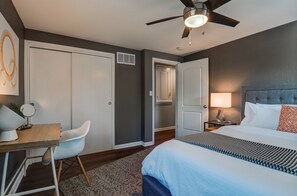 Ein Hotelzimmer mit Bett, Schreibtisch mit Stuhl, Deckenventilator, Nachttisch mit Lampe und einer Tür zu einem anderen Raum.