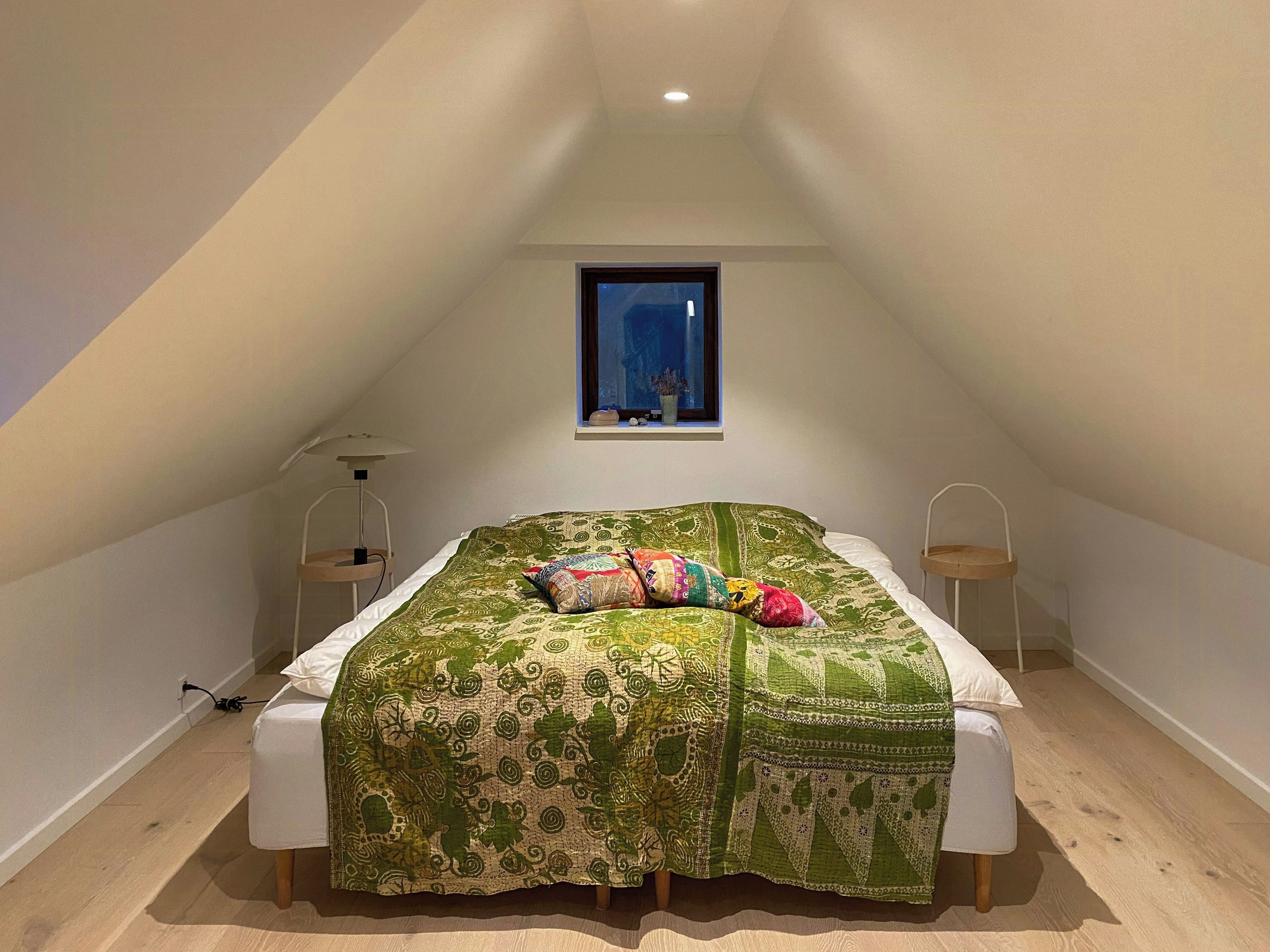 Ein Schlafzimmer mit geneigter Decke, einem Bett mit gemustertem Überwurf, zwei Nachttischen und einem Fenster mit Blick ins Freie.