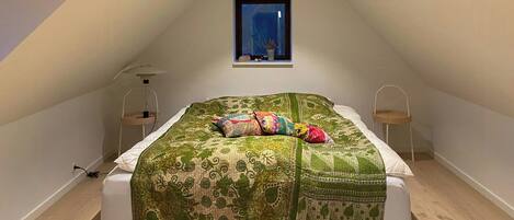 A bedroom with a sloped ceiling, a bed with a patterned bedspread, two bedside tables, and a window with a view of the outside.