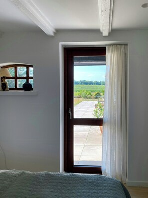 A bedroom with a bed, a window with curtains, a small shelf with a lamp and vase, and a view of a garden through the door. - Fisherman's cottage from 1873 near Bønnerup (Allingåbro)