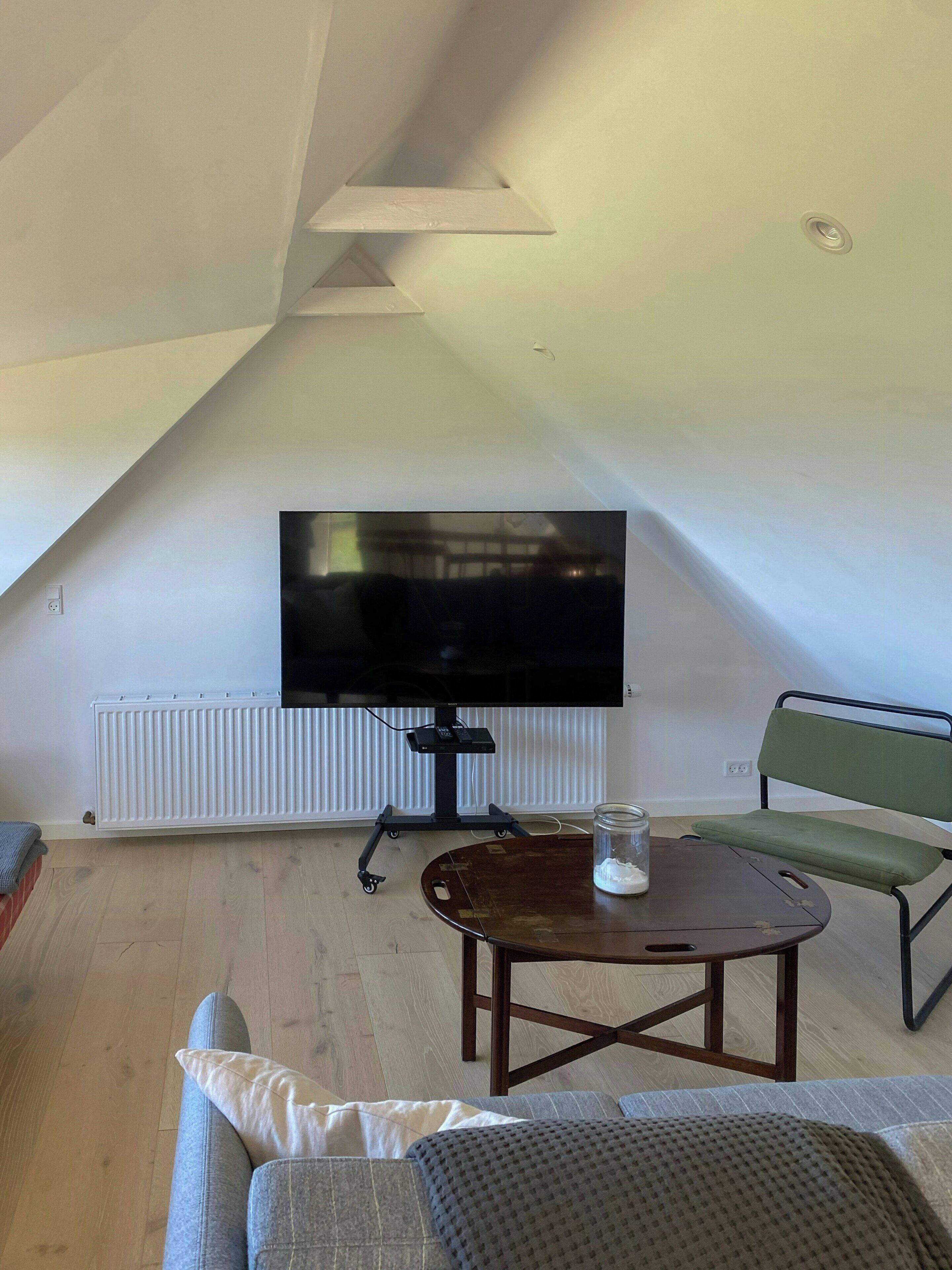 A living room with a flat-screen TV mounted on a stand, a wooden coffee table, a green chair, and a sofa with cushions.