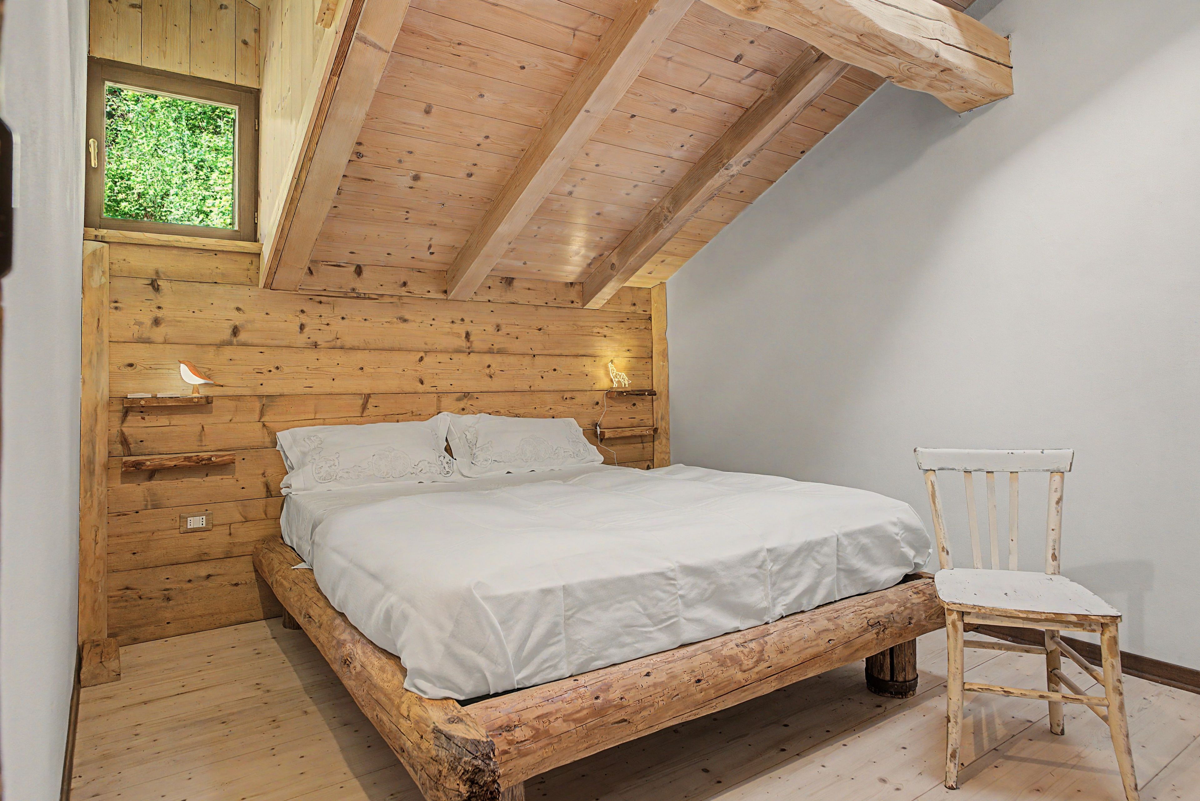 Une chambre à coucher en bois avec un plafond en pente, un lit, une table de chevet, une chaise et une fenêtre.