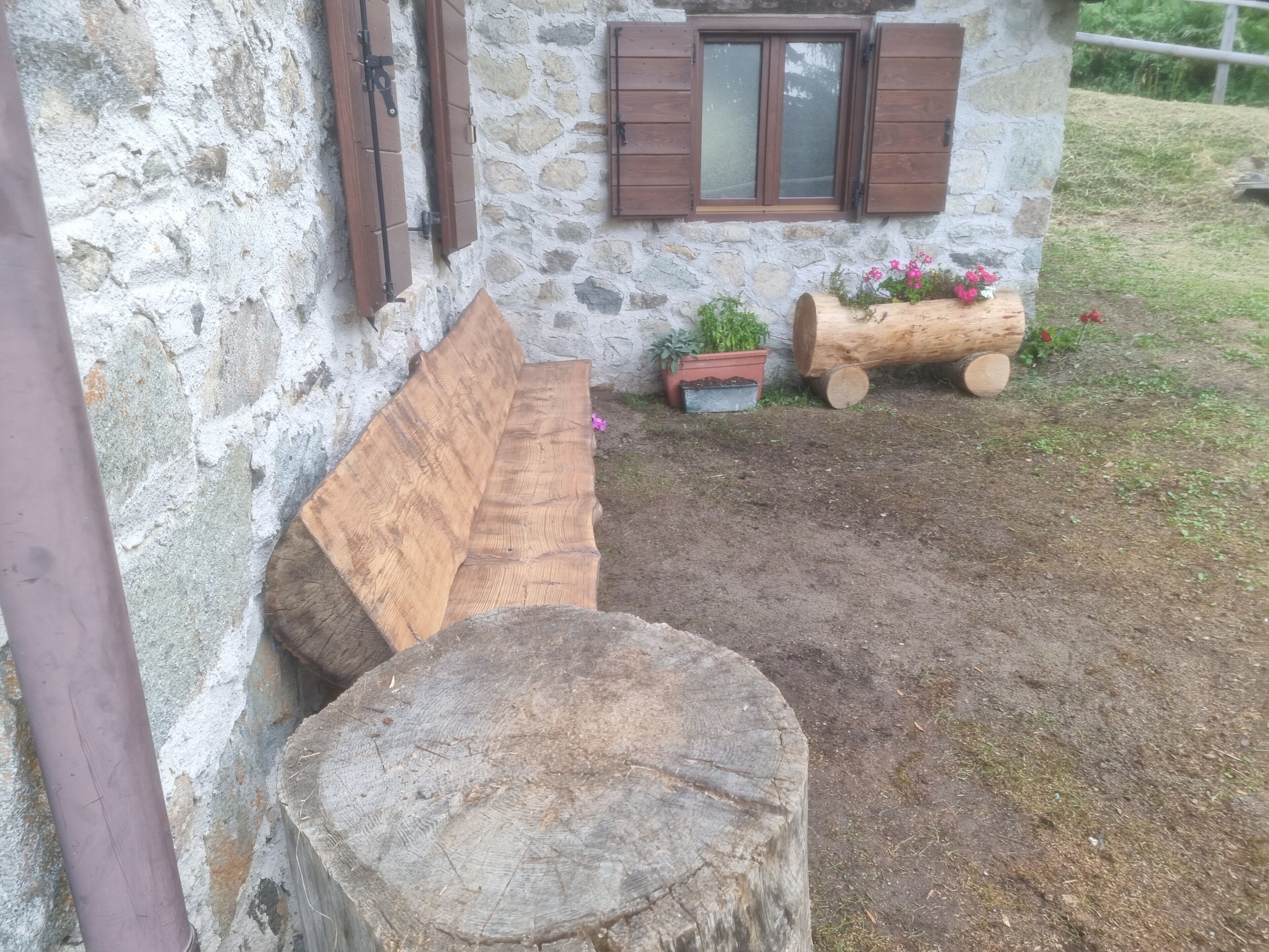 Ein rustikales Steinhaus mit Holzläden, einer Bank und einem Holzbottich als Pflanzgefäß mit Blumen.