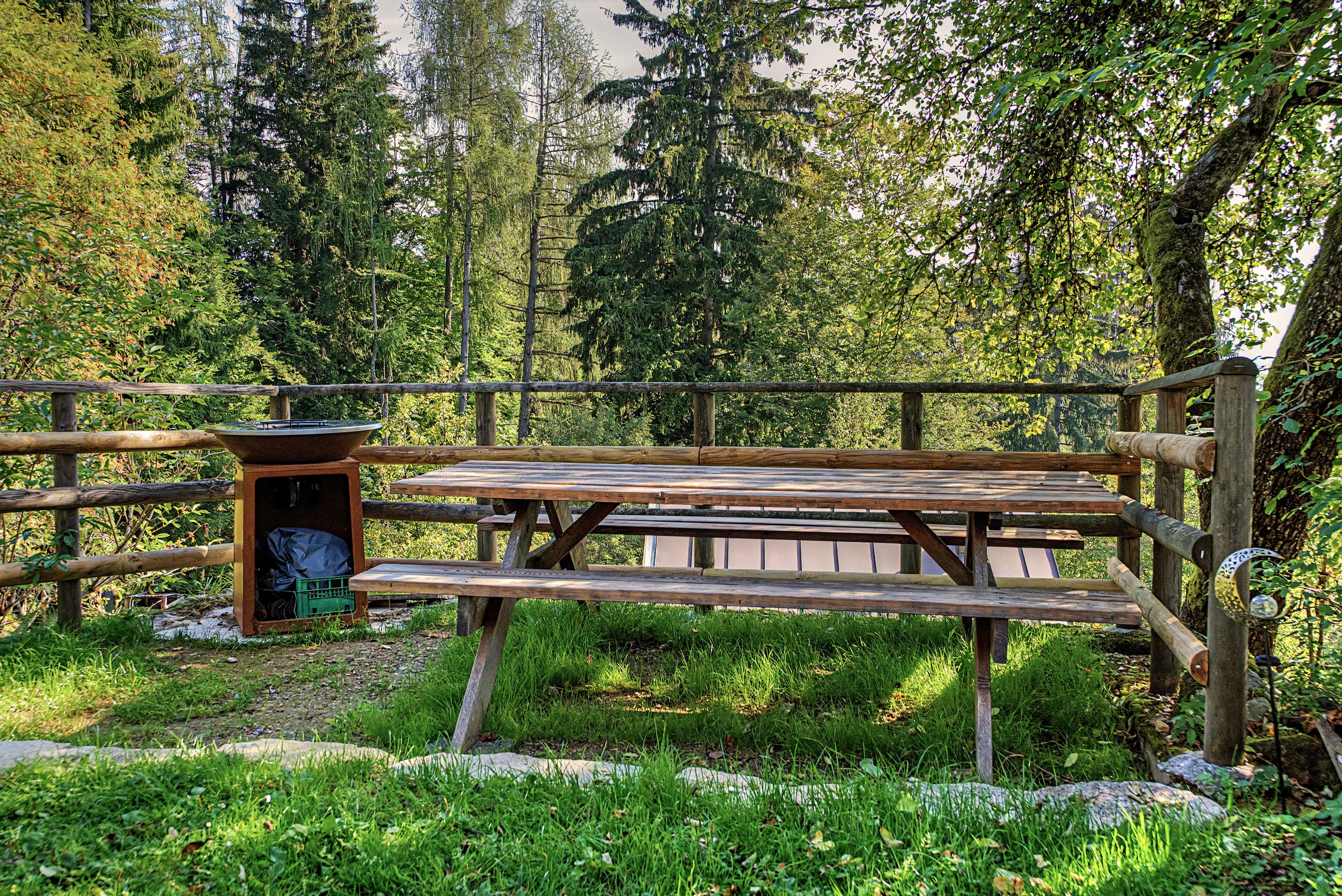 A wooden picnic table with benches, surrounded by a wooden fence, set in a forested area.