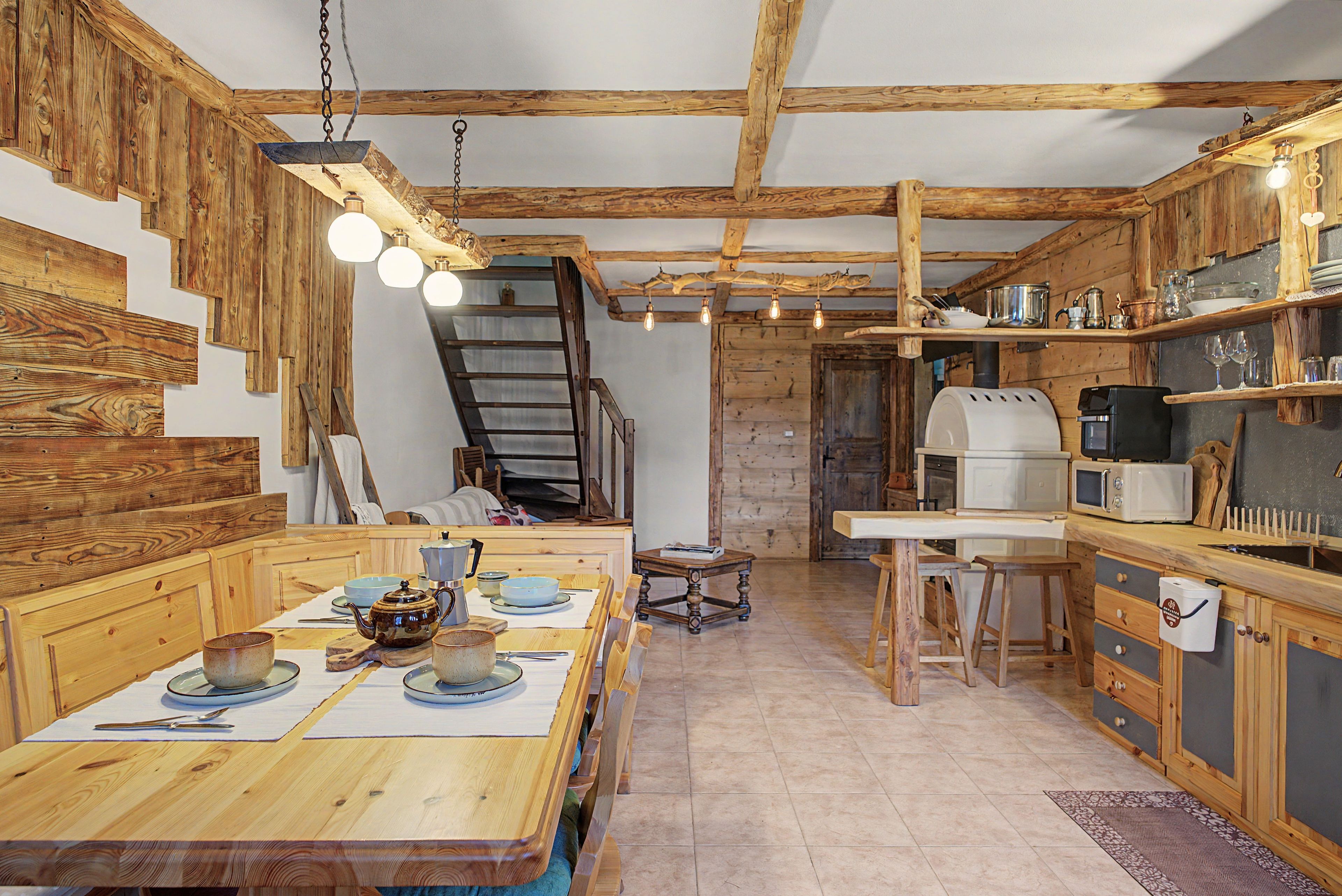 Una cocina rústica con vigas de madera, una mesa de comedor dispuesta con platos y una tetera, y una escalera que sube a un nivel superior.
