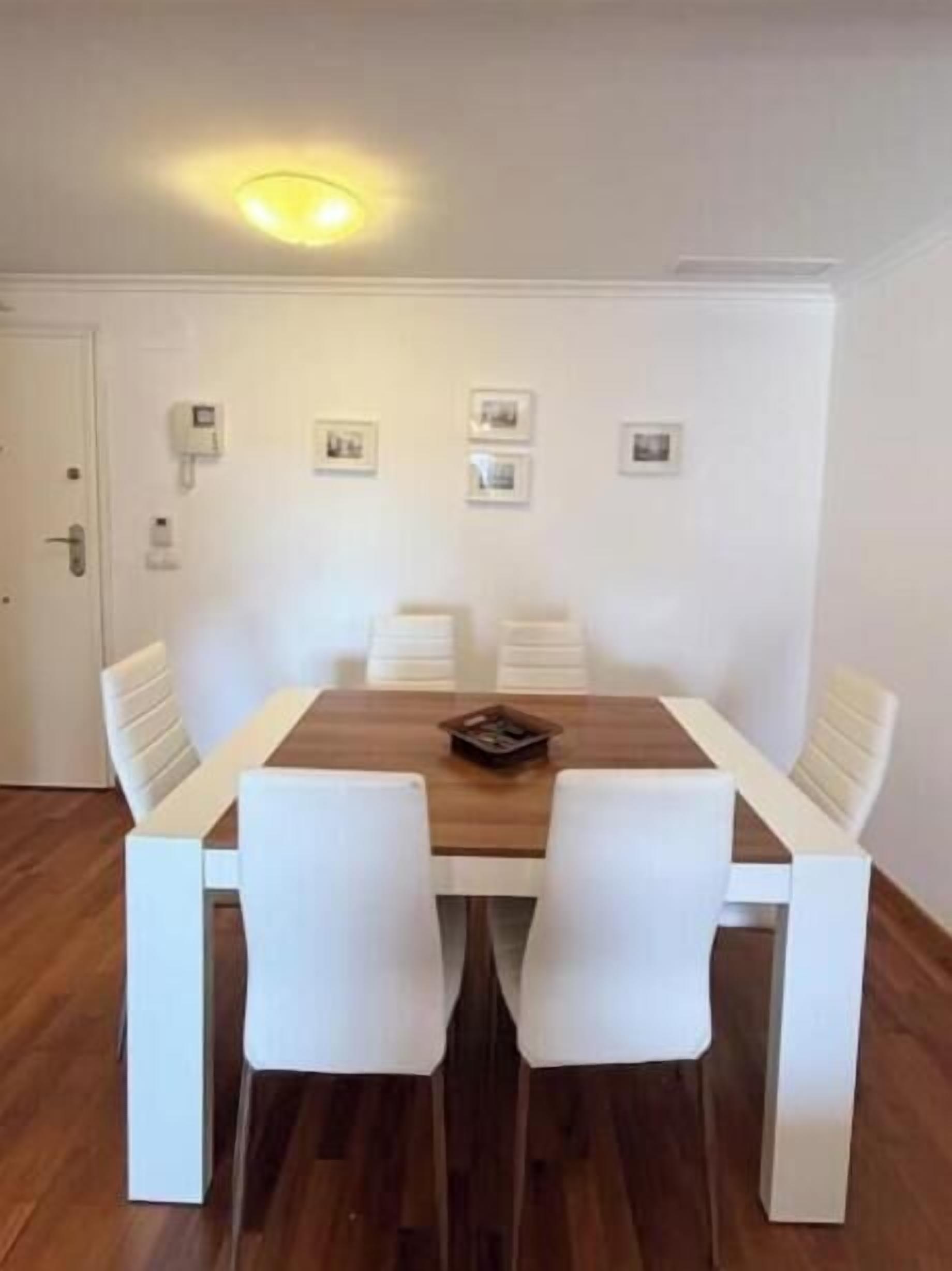 A dining area with a wooden table and white chairs, a white door, and a wall-mounted light fixture.