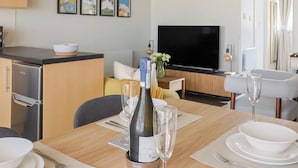 A modern kitchen with a dining area, a flat-screen TV, and a dining table set with wine glasses and a bottle. - 4 Kingfisher Court (Bridport)