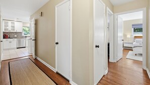 Un corridoio con due porte bianche, una zona cucina con mobili bianchi e una camera da letto con un letto e un comodino.