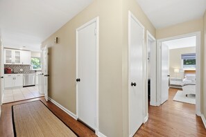 A hallway with two white doors, a kitchen area with white cabinets, and a bedroom with a bed and nightstand. - Offering the perfect balance of quiet neighborhood living and urban convenience. (Durham)