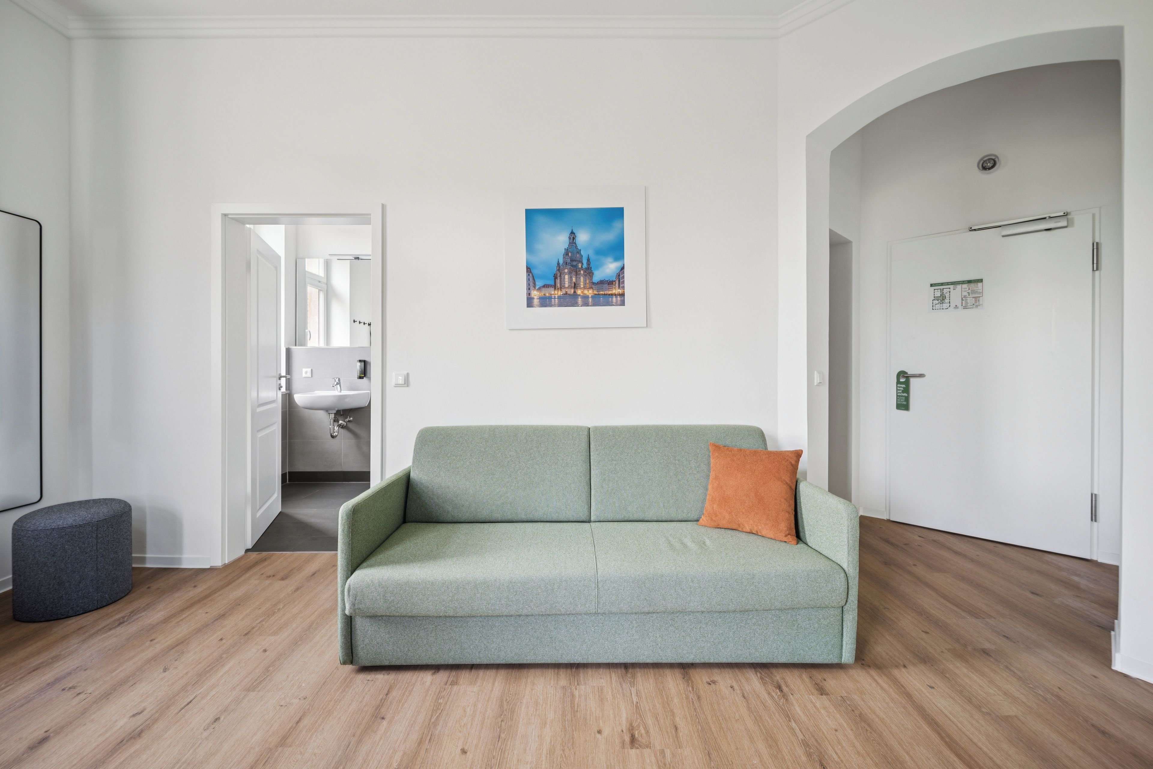 Ein Zimmer mit einem grünen Sofa, einem kleinen runden Hocker, einem Bild eines Gebäudes an der Wand und einer Tür mit einem Schild.