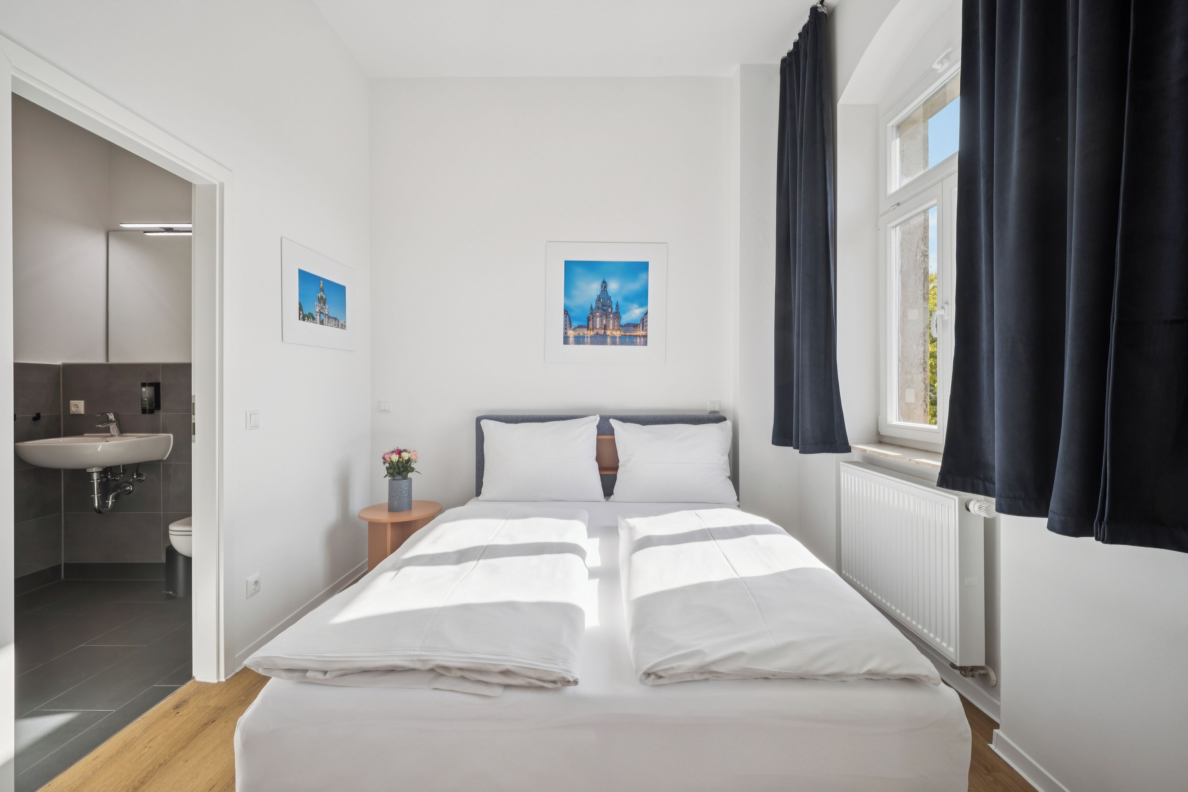Ein Hotelzimmer mit Bett, einem Fenster mit Vorhängen, einem Nachttisch mit Blumenvase und zwei gerahmten Bildern an der Wand.