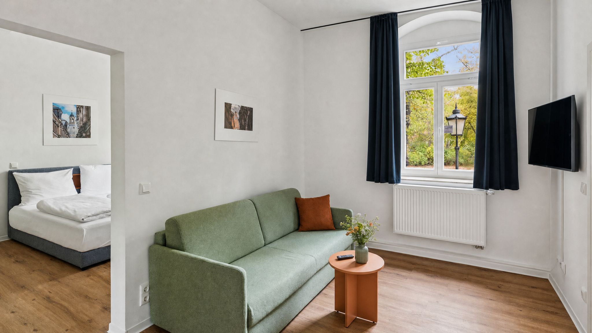A room with a green sofa, a bed with white bedding, a small round table with a vase, and a TV mounted on the wall.