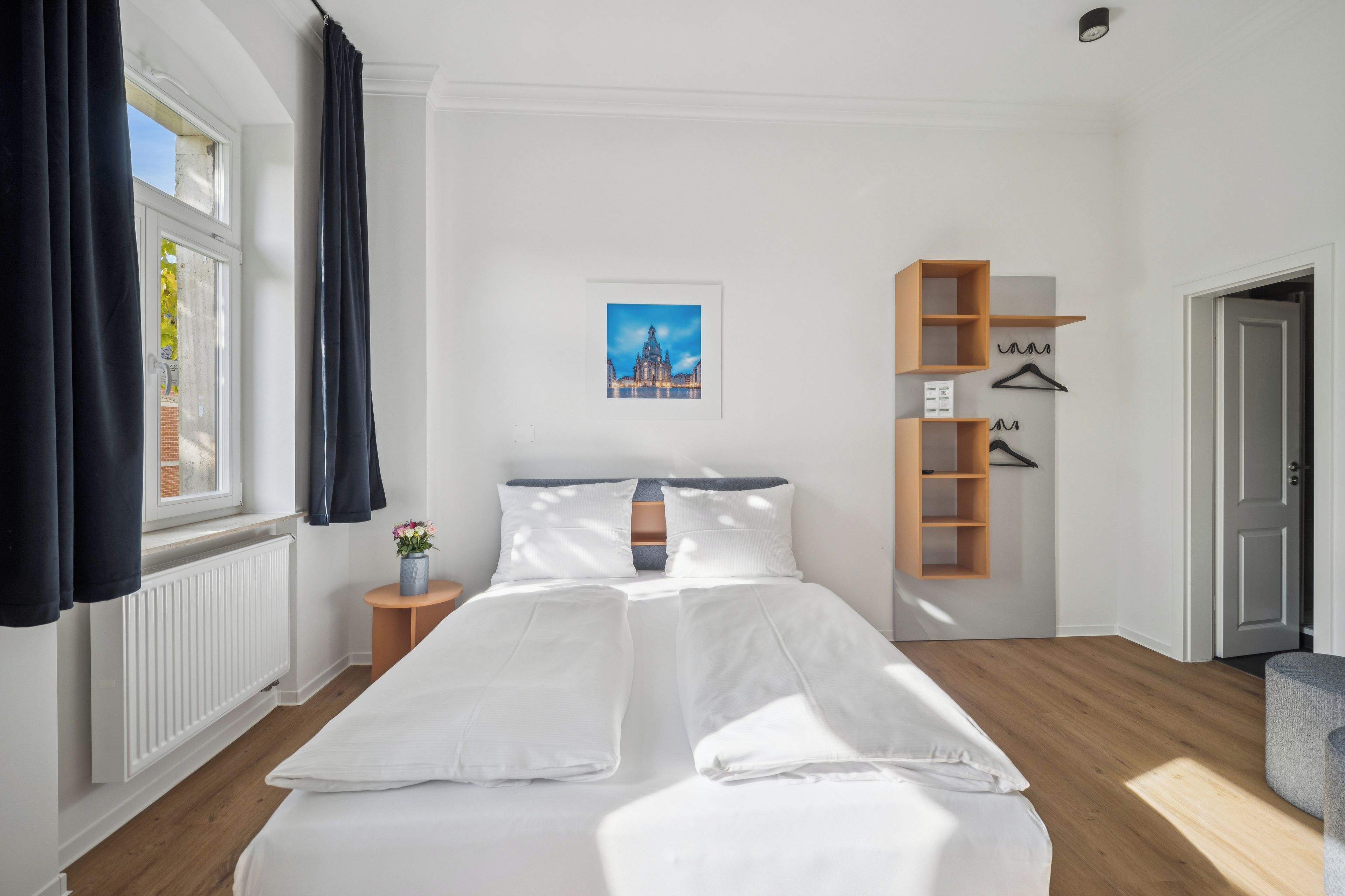 Une chambre d’hôtel avec un grand lit, une table de chevet ornée d’un vase de fleurs, un tableau encadré au mur et une étagère en bois avec des cintres.