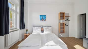 Habitación de hotel con una cama grande, una mesita de noche con un florero, un cuadro en la pared y un estante de madera con perchas.
