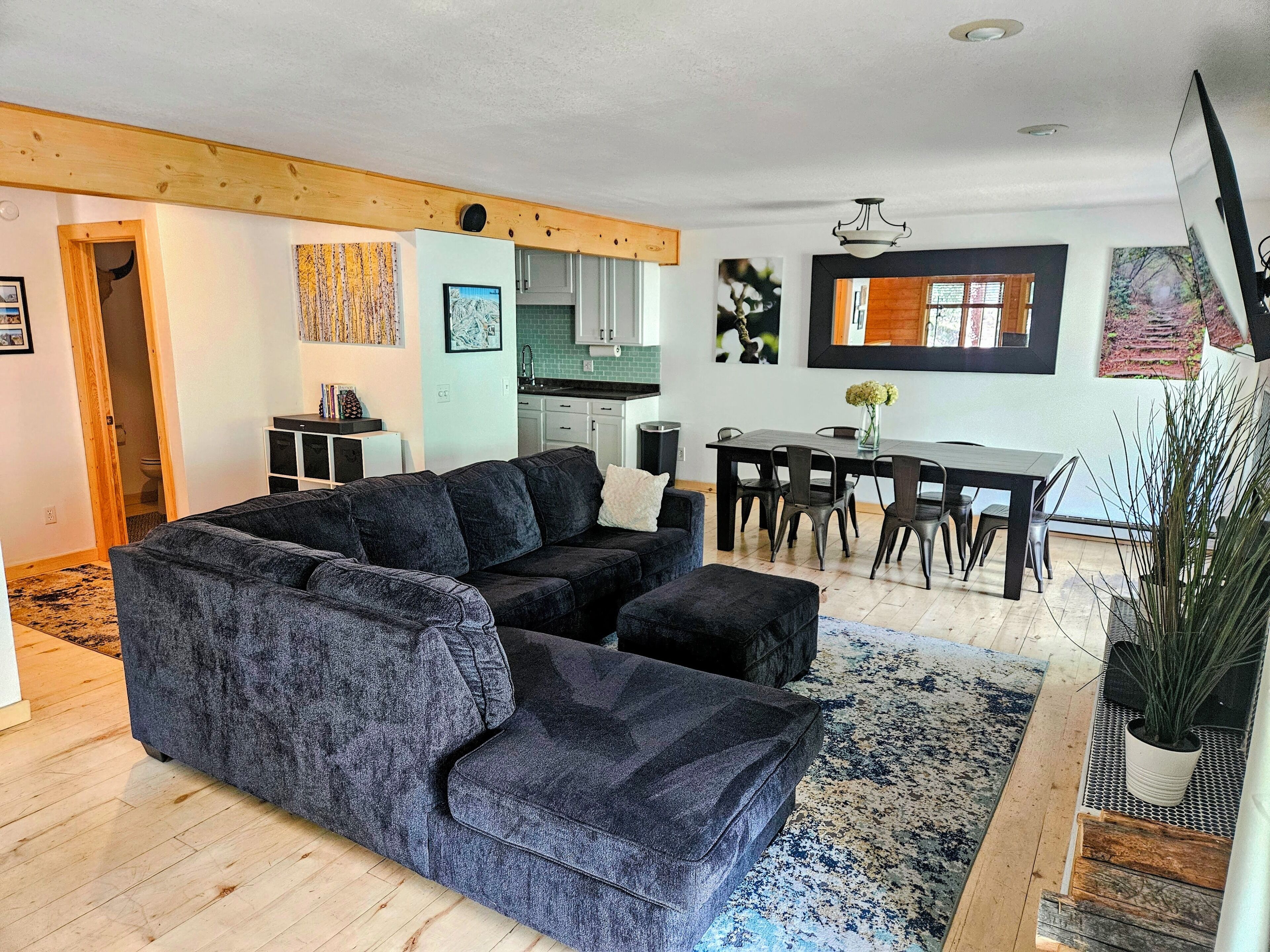 Een moderne woonkamer met een hoekbank, een eettafel met stoelen, een keuken op de achtergrond en een trap die naar een andere verdieping leidt.