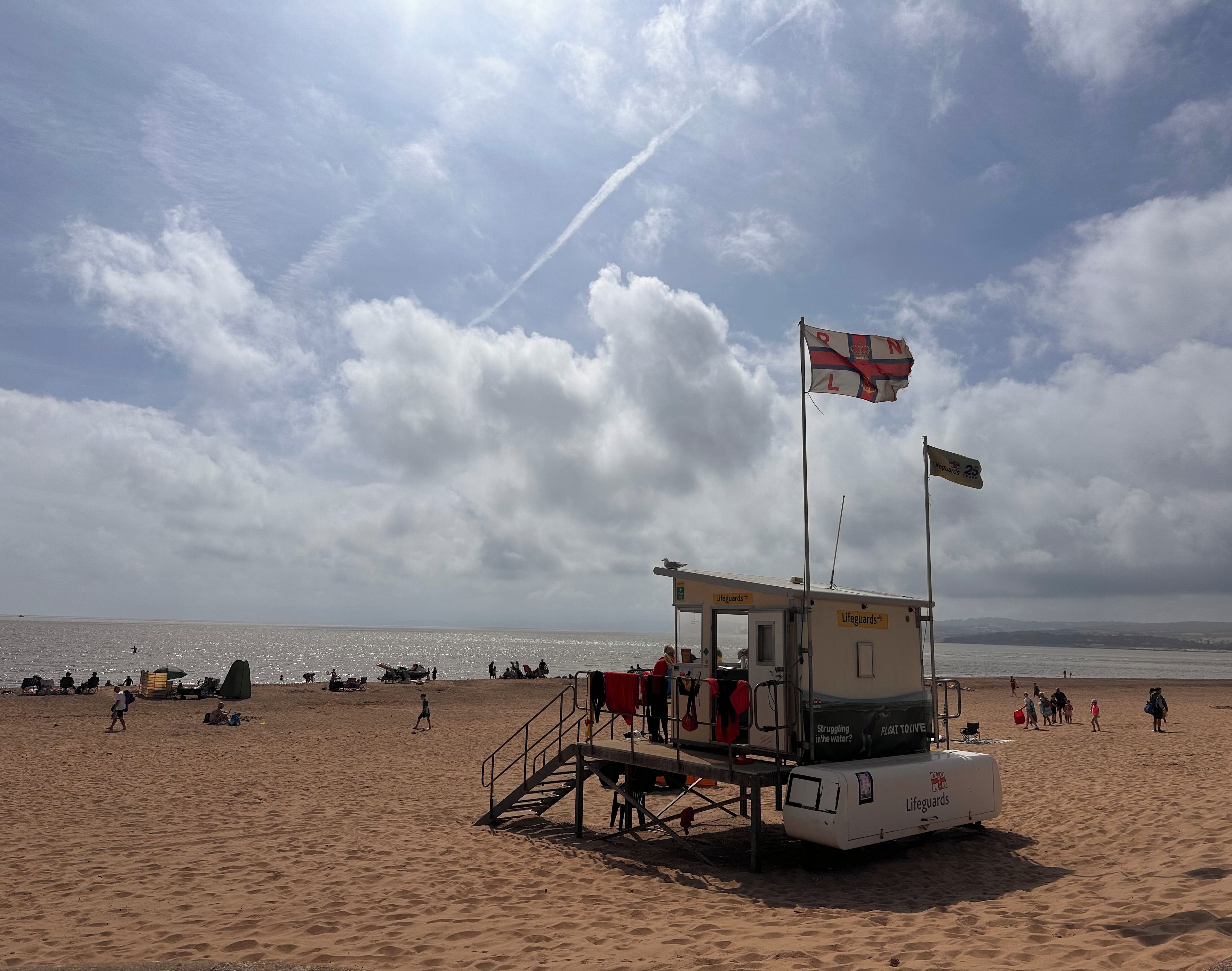 A lifeguard station on a beach with a flag, a lifeguard booth, and people in the background.