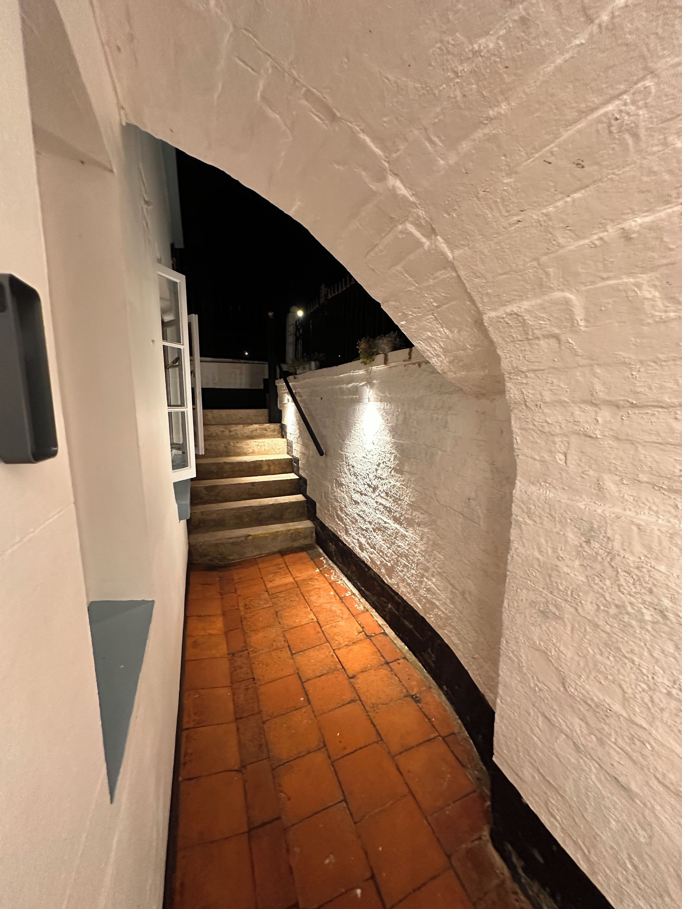 A narrow, brick-paved corridor with a staircase leading upwards, flanked by white walls and a single illuminated light fixture.