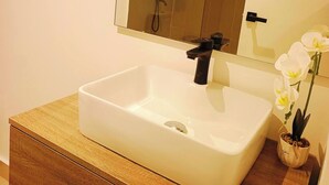 Een badkamer met een wit wastafelmeubel, een houten kastje, een spiegel en een douche die op de achtergrond zichtbaar is.