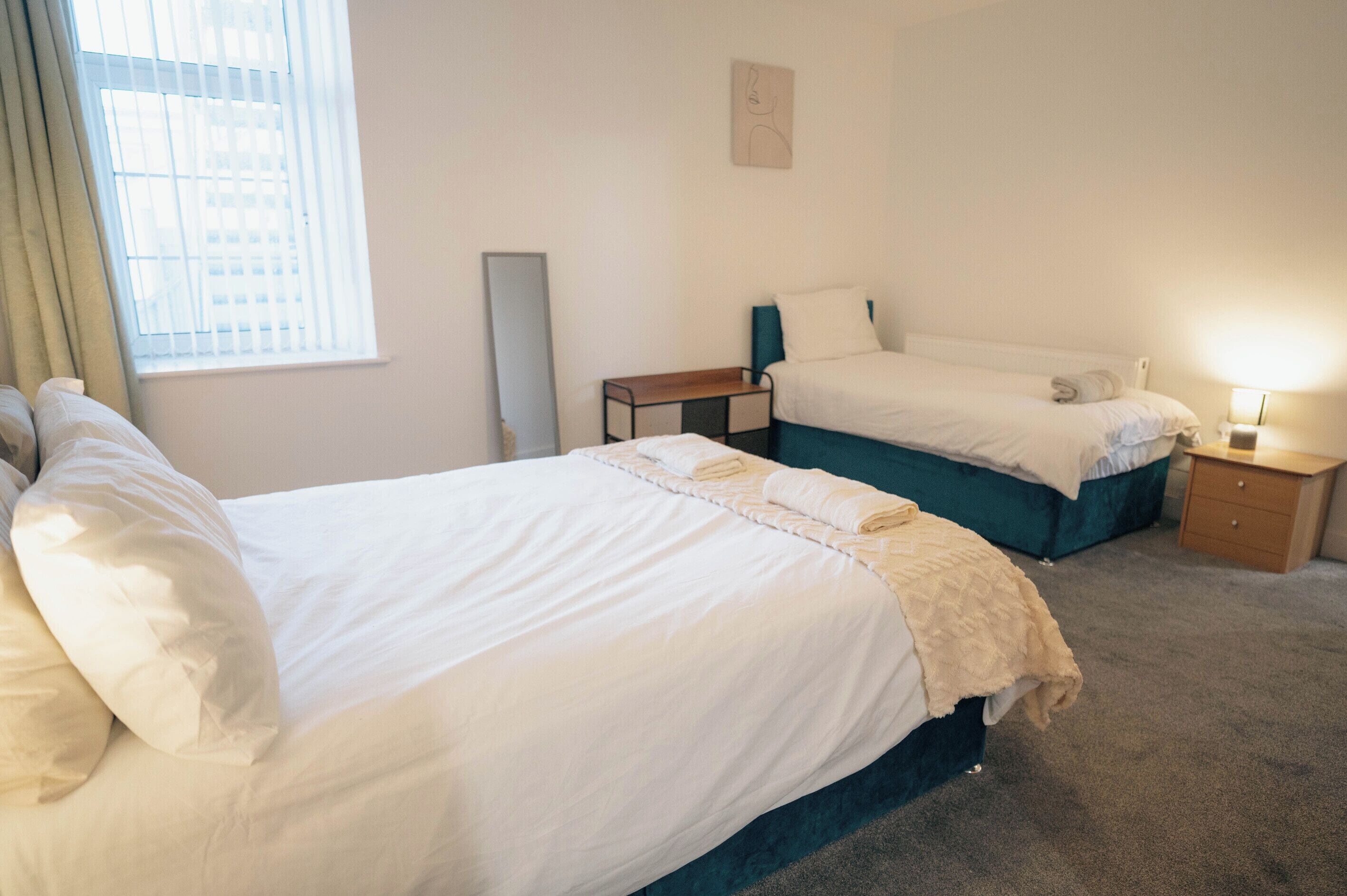 A hotel room with two beds, a bedside table, a lamp, and a window with blinds.