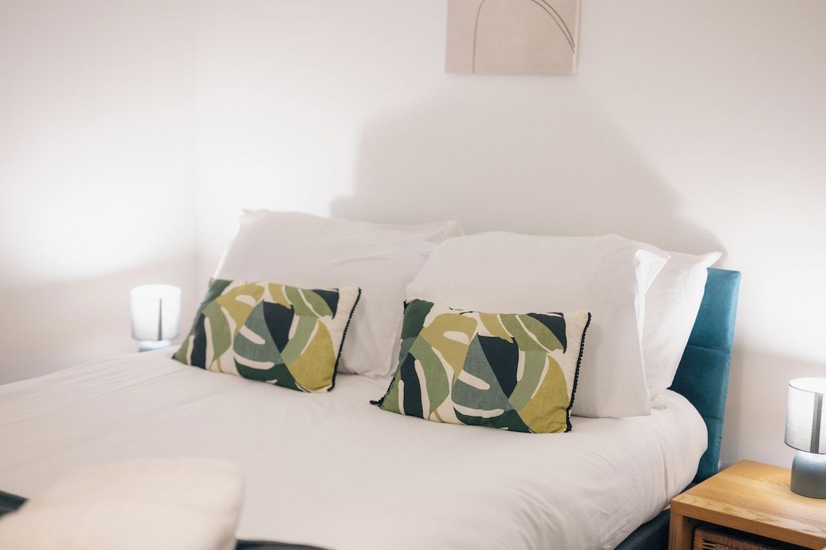 Ein Bett mit weißer Bettwäsche und zwei gemusterten Kissen, ein Nachttisch mit Lampe sowie ein an der Wand hängendes Kunstwerk.