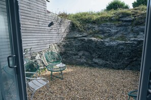 Un patio con dos sillas verdes y una mesa, suelo de grava y una pared de madera.