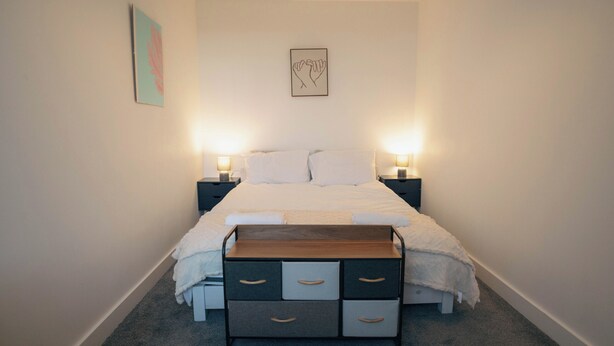 A bedroom with a bed, bedside tables, a nightstand, a wall-mounted artwork, and a carpeted floor. - Beach Court by Guestz (Trearddur Bay)