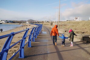 Ein Küstenweg mit blauer Geländer, ein Strand und Häuser im Hintergrund.