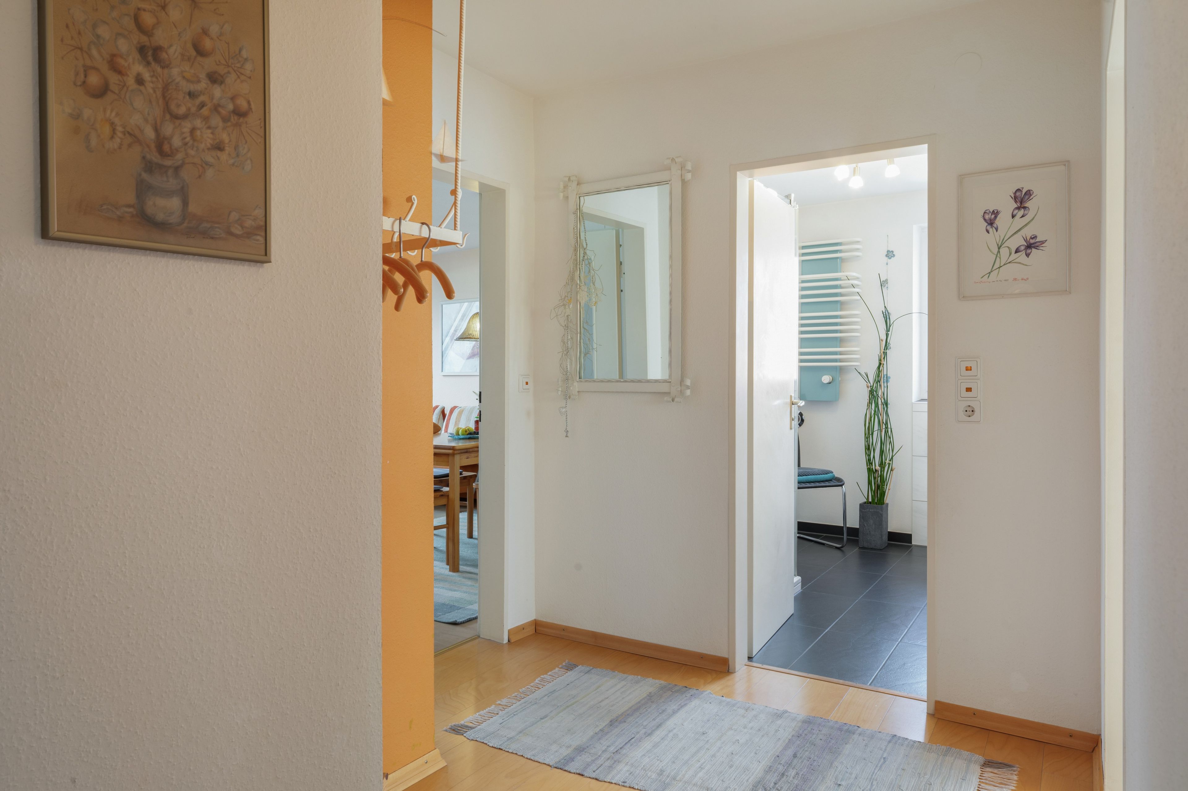 A hallway with a wooden floor, a rug, a mirror, and a door leading to another room.