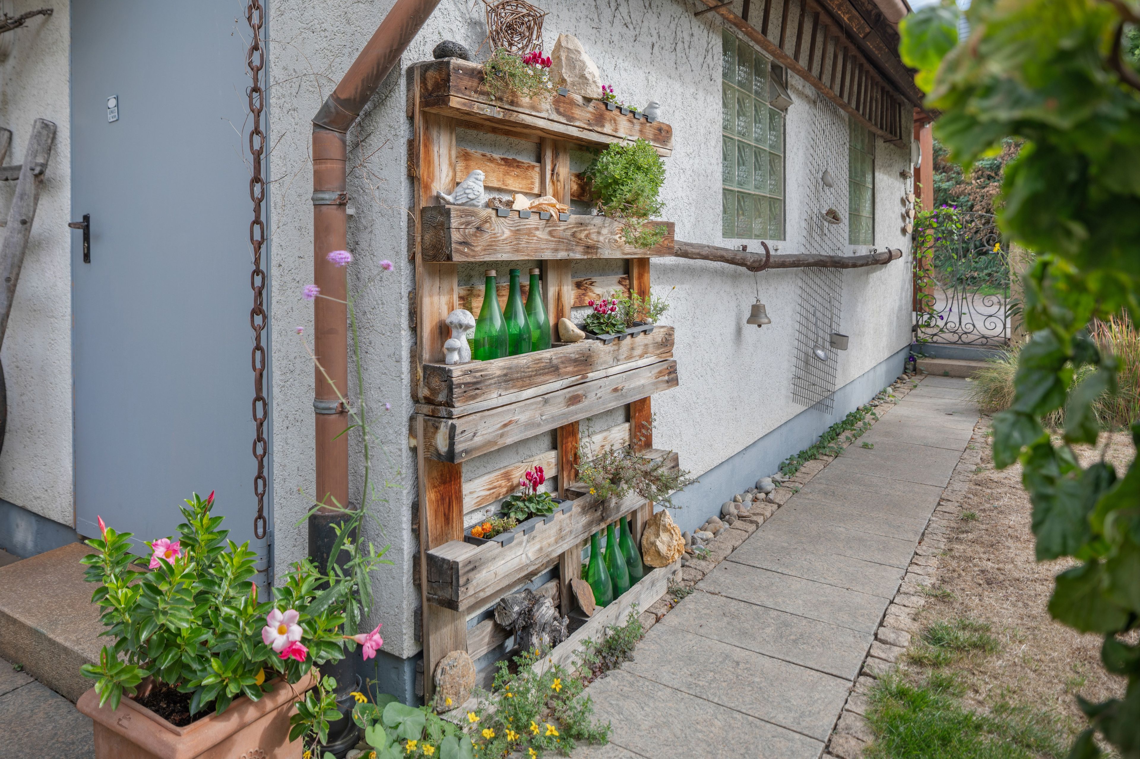 Un edificio rústico con una estantería de madera adosada a la pared exterior, mostrando diversos objetos como botellas, flores y adornos.