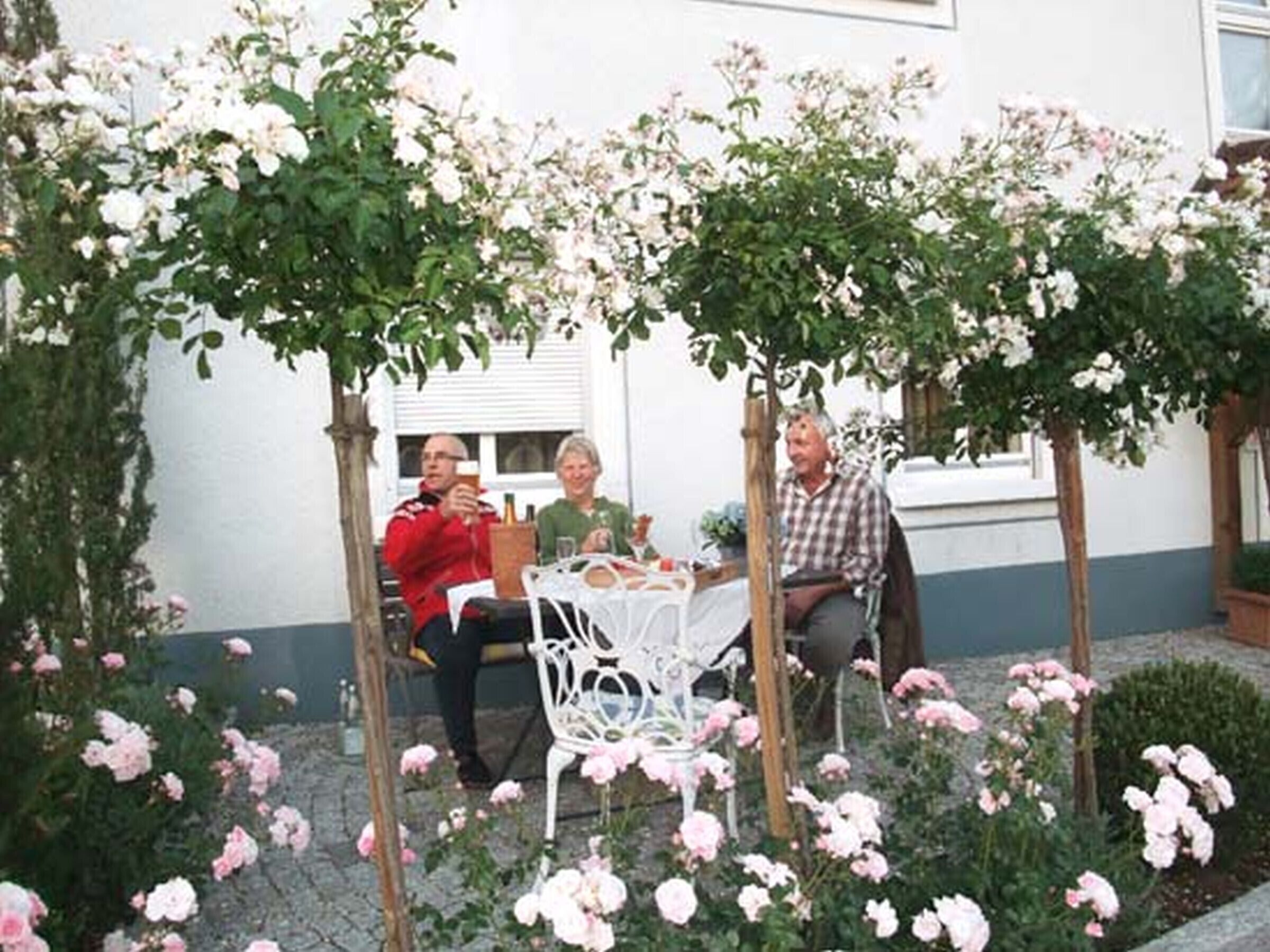 A patio area with a table and chairs, surrounded by flowering plants and a white fence.
