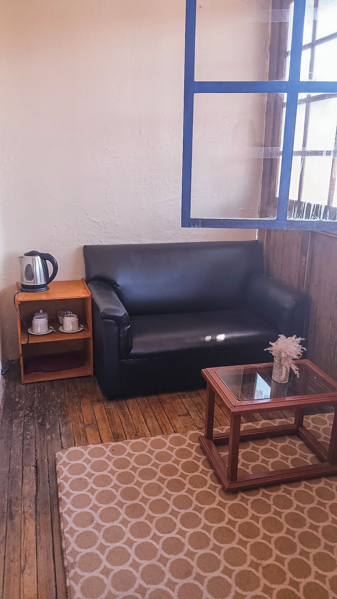 A room with a black leather sofa, a wooden side table with a kettle and cups, a glass coffee table with a vase, and a patterned rug.