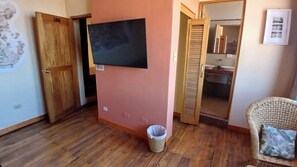 A room with a wooden floor, a television mounted on the wall, a wicker chair with a cushion, and a door leading to a bathroom. - Loki Boutique Cusco (Cusco)