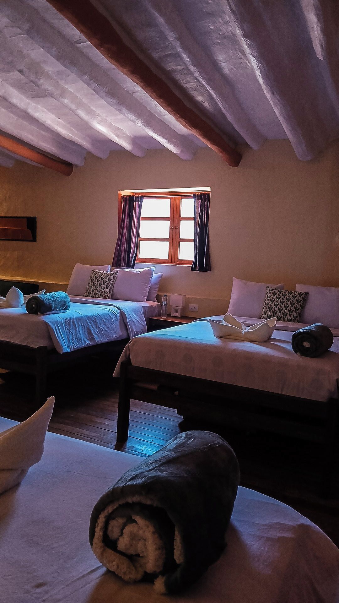 Habitación de hotel con dos camas, una ventana con cortinas y techo de madera.