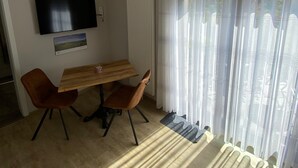 Un coin repas avec une table et des chaises, un téléviseur à écran plat fixé au mur, un radiateur et une fenêtre ornée de rideaux légers.