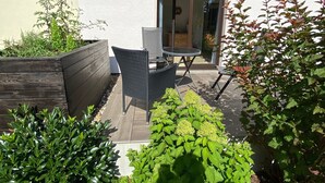 Une terrasse avec une table et des chaises, entourée de différentes plantes et d’un jardinier en bois.