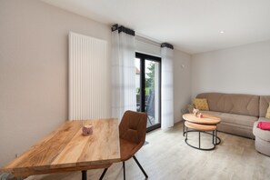 A modern living room with a wooden dining table, a brown sofa, and a small round table with a vase. - Holiday Apartment "Auszeit" with Private Terrace, Garden, and Wi-Fi (Bad Liebenwerda)