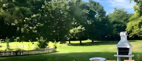 Un área de estar al aire libre con una parrilla, rodeada de árboles y un campo de césped.
