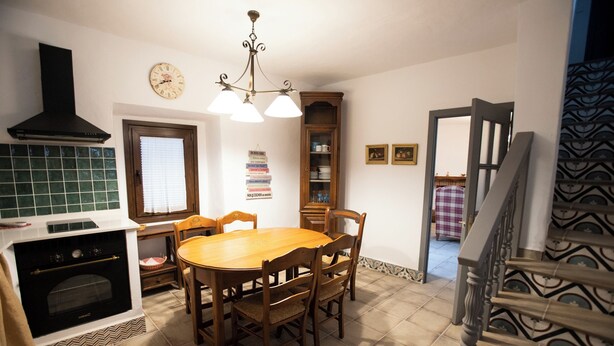 A kitchen with a dining table, chairs, a clock, a cabinet, and a staircase. - Casa rural La Bandolera (Grazalema)