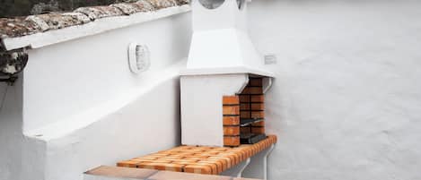 Ein kleines Kochareal im Freien mit einem Backofen aus Ziegeln, einer weißen Wand und einer hölzernen Bank.