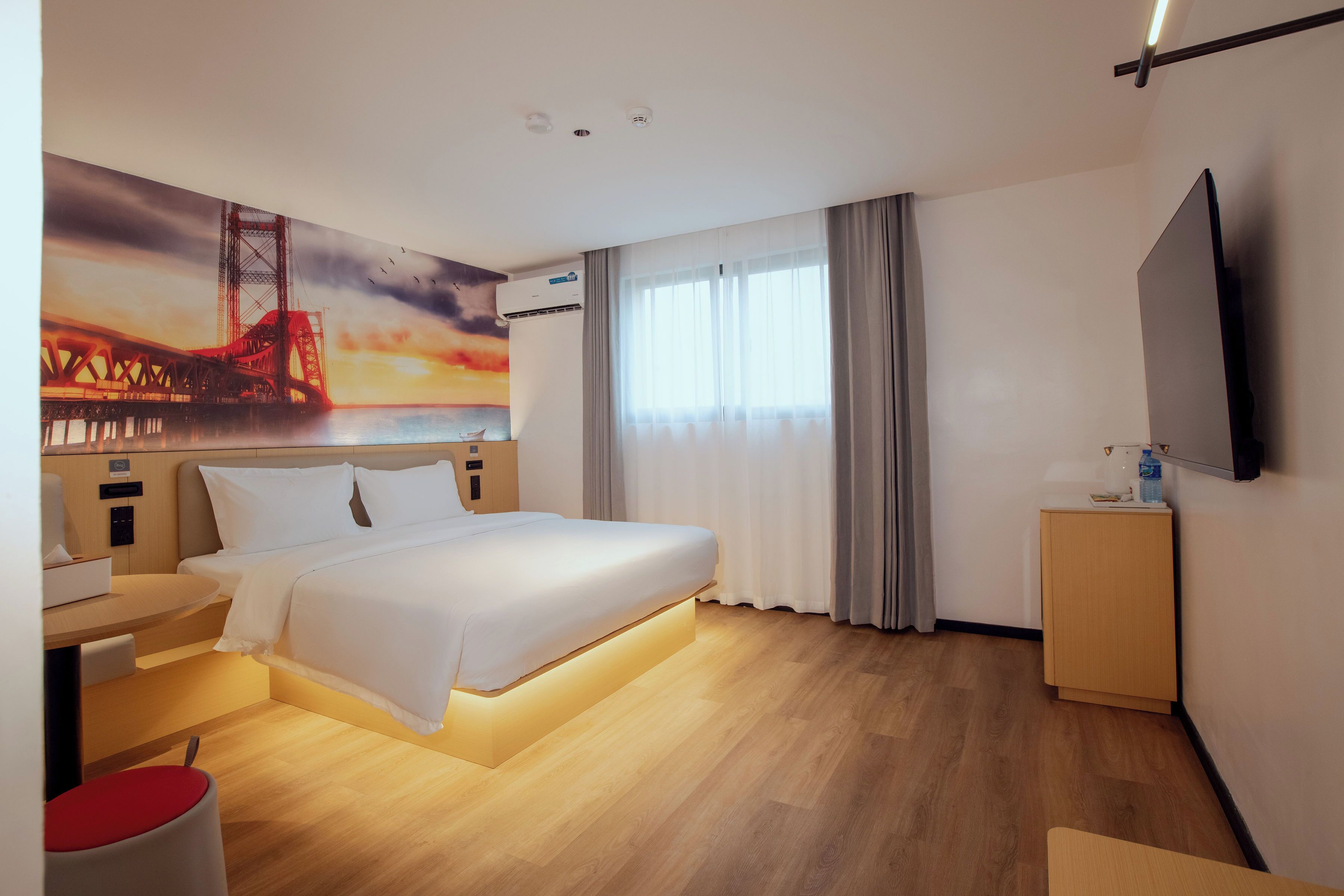 A hotel room with a large bed, a desk, a chair, a TV, and a painting of a bridge.