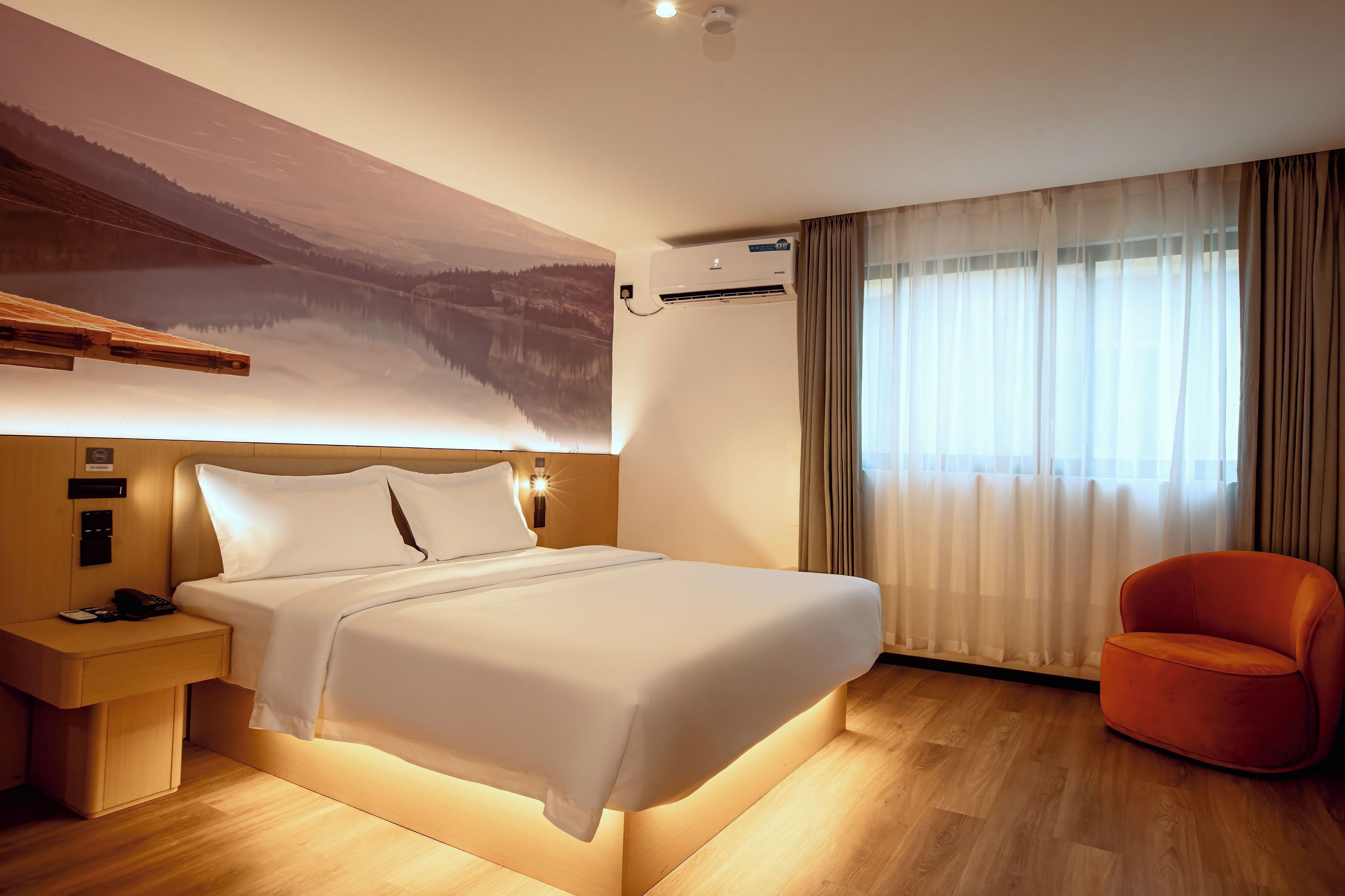A hotel room with a large bed, a nightstand, a red chair, and a wall-mounted air conditioning unit.