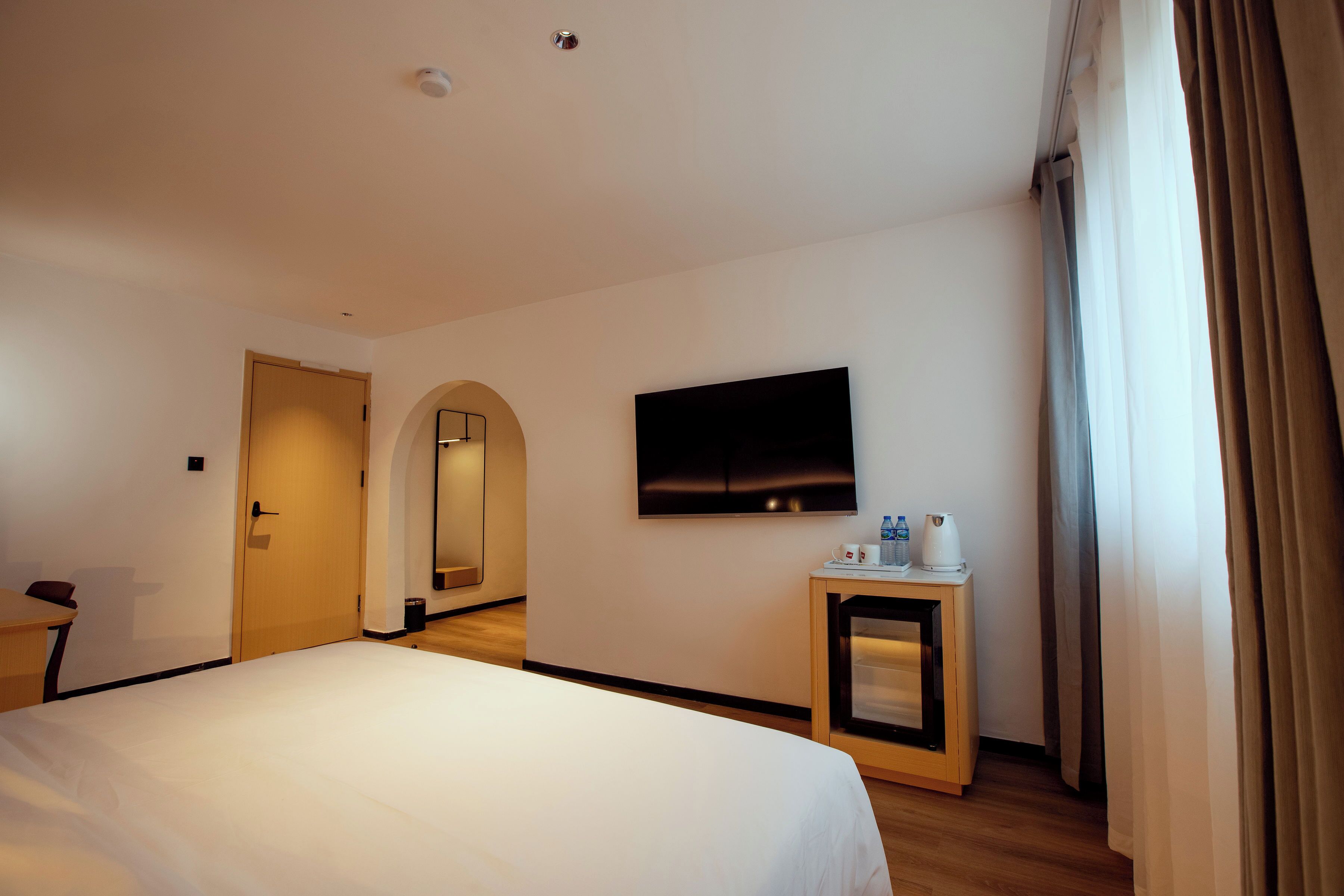 Hotelkamer met een bed, een aan de muur bevestigde televisie, een klein tafeltje met waterkoker en kopjes, en een deur naar een andere kamer.