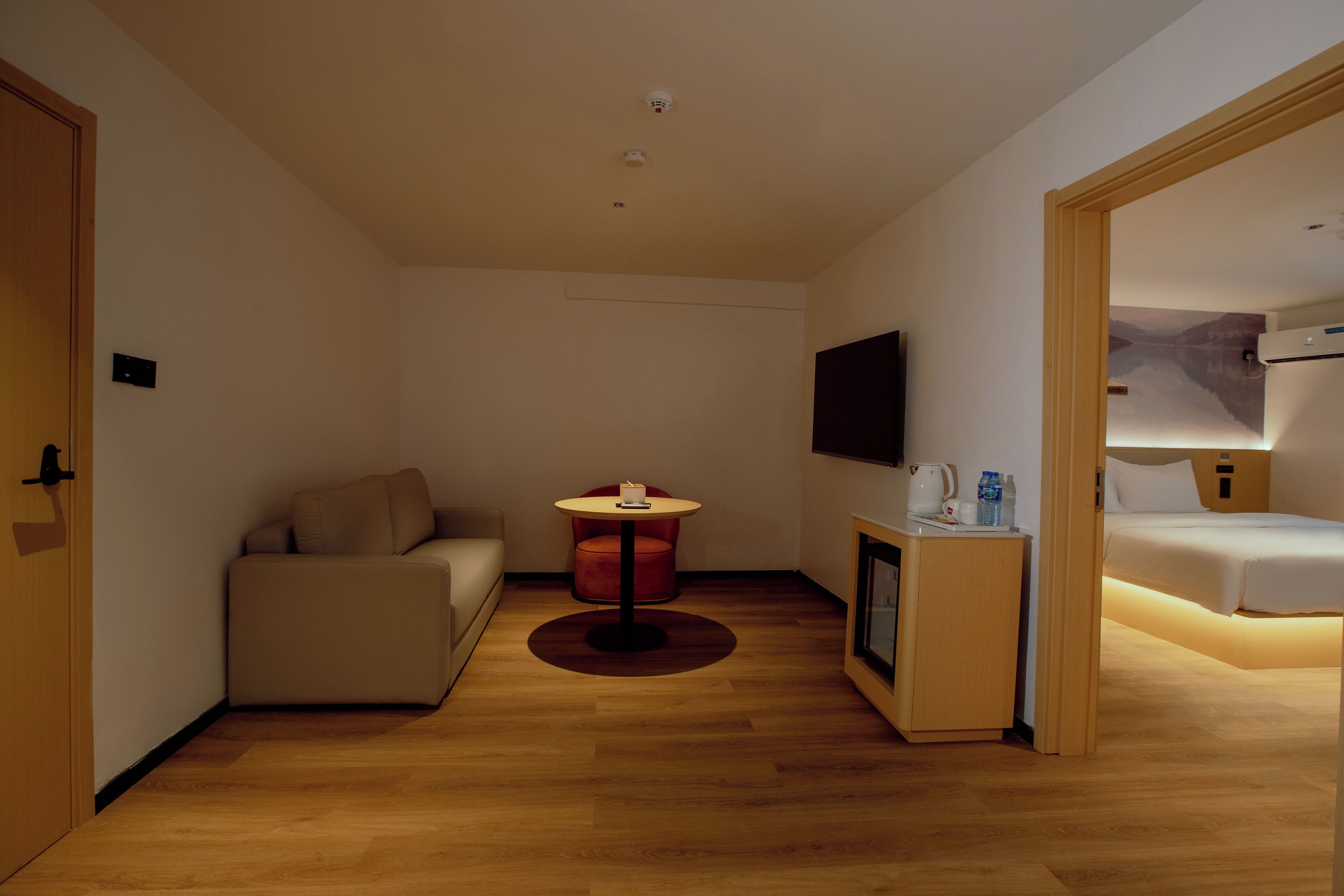 Habitación de hotel con cama, sofá, una mesita con lámpara, televisor montado en la pared y suelo de madera.