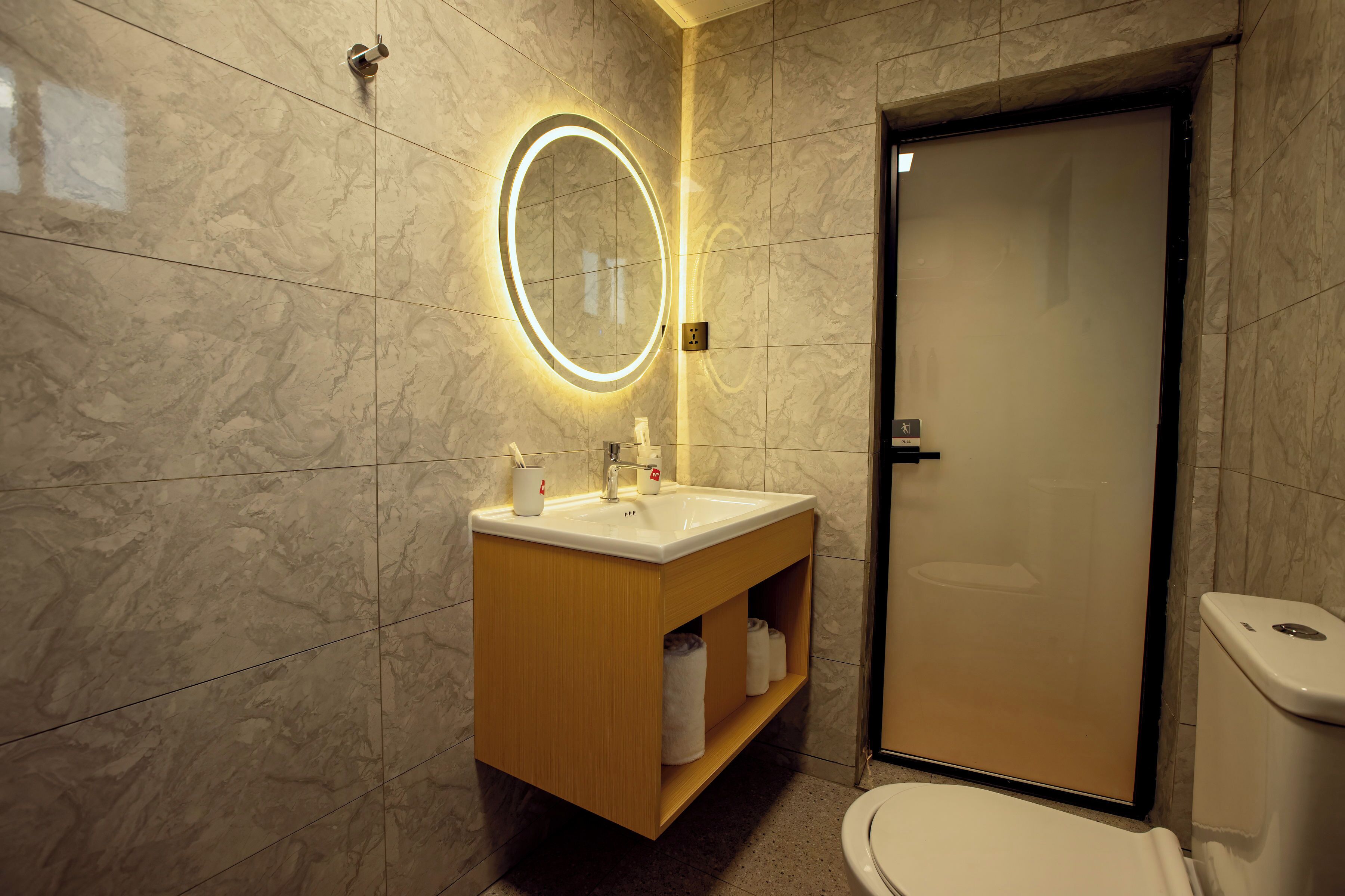 Un baño con pared de mármol, tocador de madera con lavamanos, espejo con luces, inodoro y una puerta.