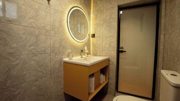 Un baño con pared de mármol, tocador de madera con lavamanos, espejo con luces, inodoro y una puerta.