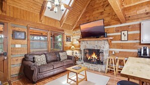 Interior acolhedor de uma cabana com um sofá de couro, lareira de pedra, televisão e paredes de madeira.