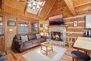 A cozy cabin interior with a leather sofa, a stone fireplace, a television, and wooden walls. - Cabin Fever by Beyond Expectations: 5-Star Cabin w/ Hot Tub, FirePit & Game Room (Gatlinburg)