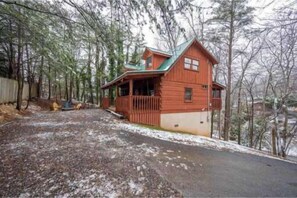 A wooden cabin with a green roof, surrounded by trees, and a paved driveway. - Cabin Fever by Beyond Expectations: 5-Star Cabin w/ Hot Tub, FirePit & Game Room (Gatlinburg)