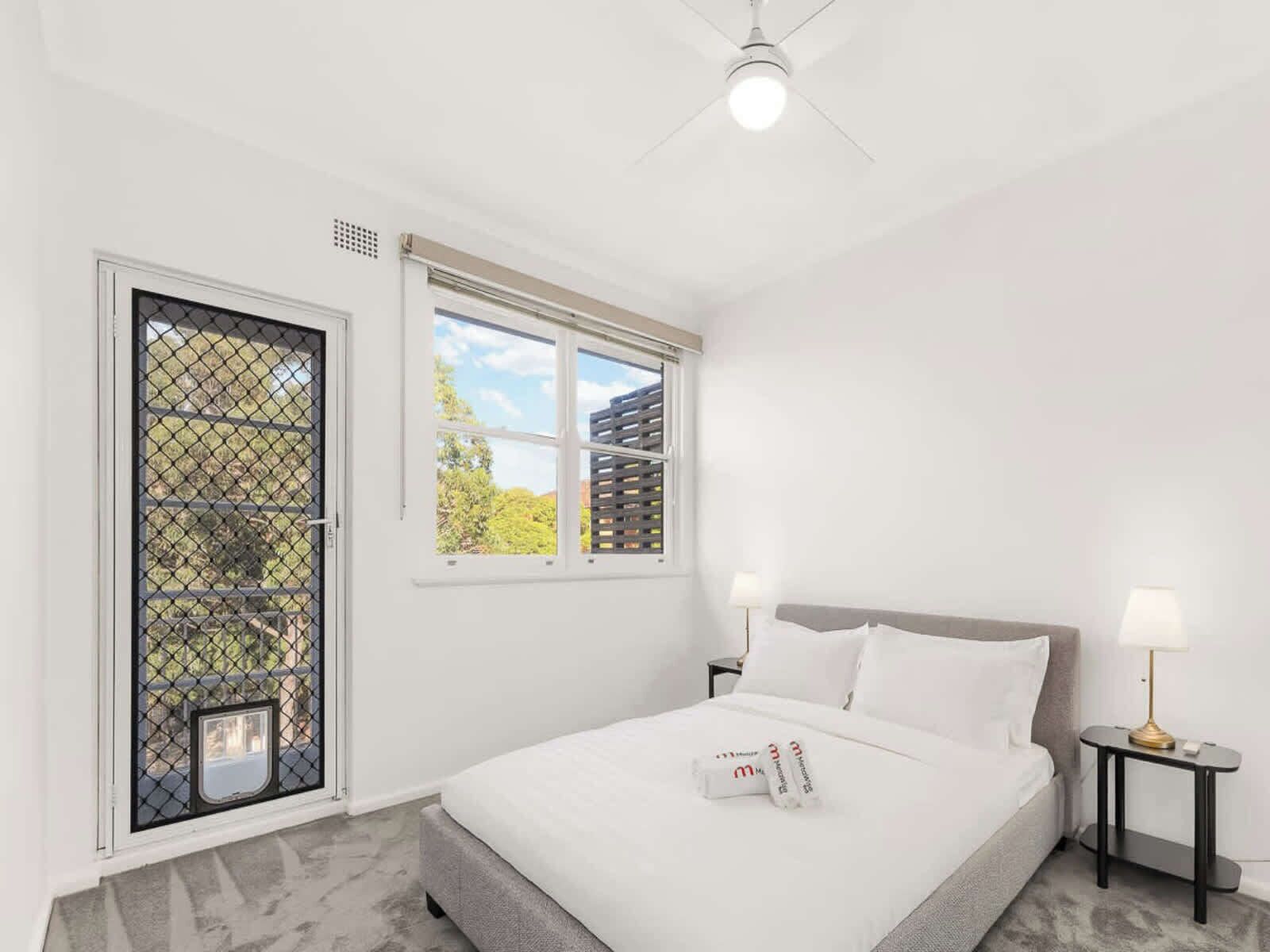 A bedroom with a bed, two bedside tables with lamps, a window with blinds, and a door with a security screen.